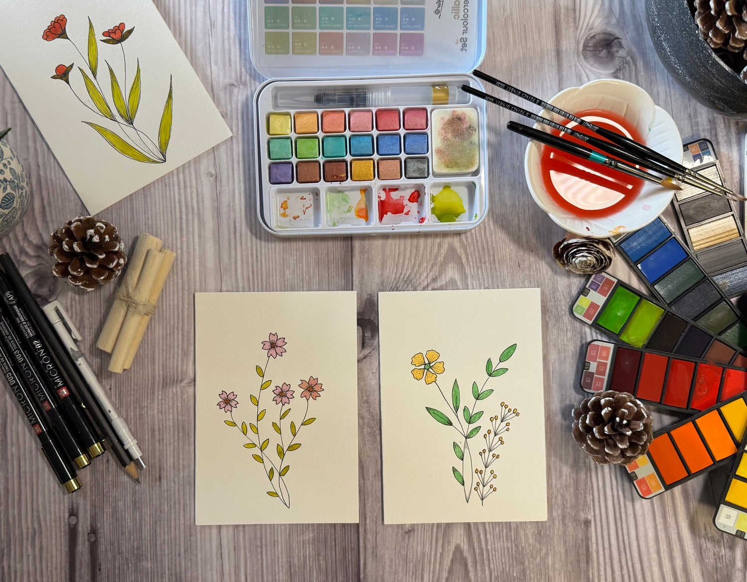 Hand painted cards placed on a table, sakura pens, watercolour paints, paint brushes and few decorative itesm placed around the cards