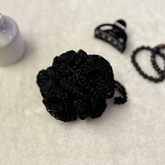 Black hand crocheted loofha with a hang tie placed on a soft fabric with trinkets in the background
