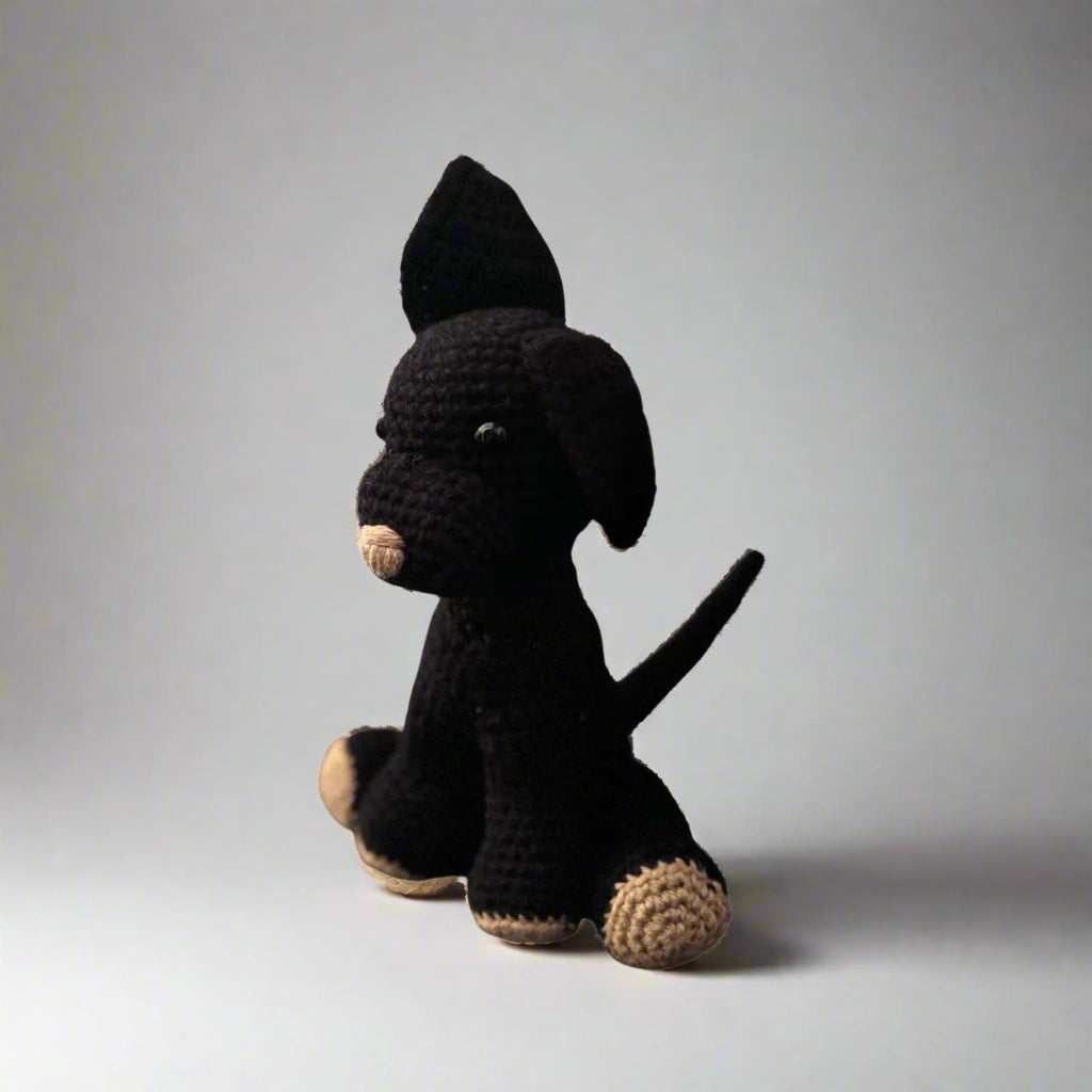 Black hand crocheted dog sitting with the hind paws spread out and the front paws placed in front with the tail upright and one ear up in the air