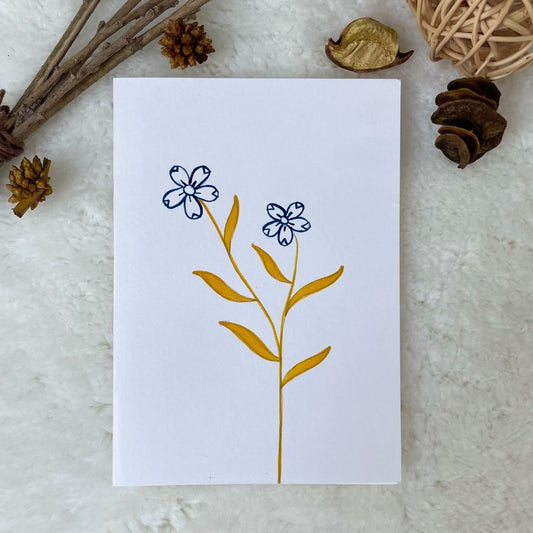 A hand painted greeting card with various flower stalks painted on a white card placed on a a fur cloth with decorative flowers in the background