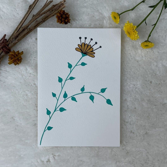 A hand painted greeting card with a yellow flower stalk painted on a white card placed on a a fur cloth with decorative flowers in the background