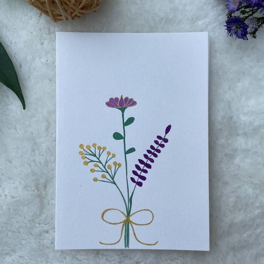 A hand painted greeting card with various flower stalks painted on a white card placed on a a fur cloth with decorative flowers in the background