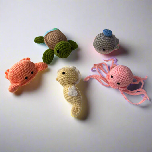 Crochet plushie set of a orange crab, yellow seahorse, grey whale, green turtle and pink octopus placed on a w white table