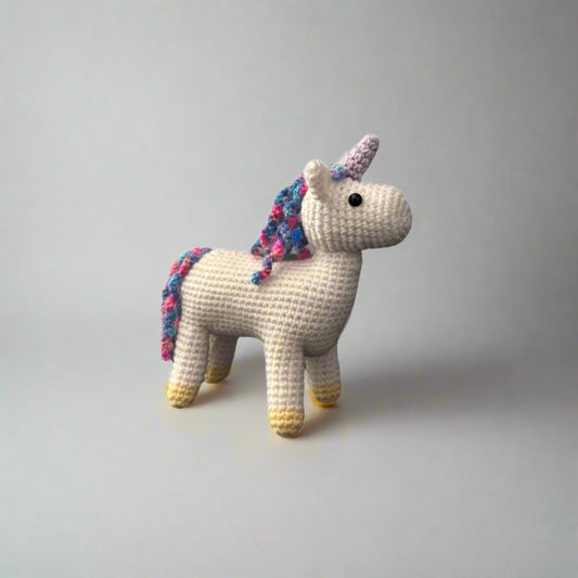 Crocheted white unicorn plushie with a purple horn and rainbow coloured mane and tail standing on a white surface