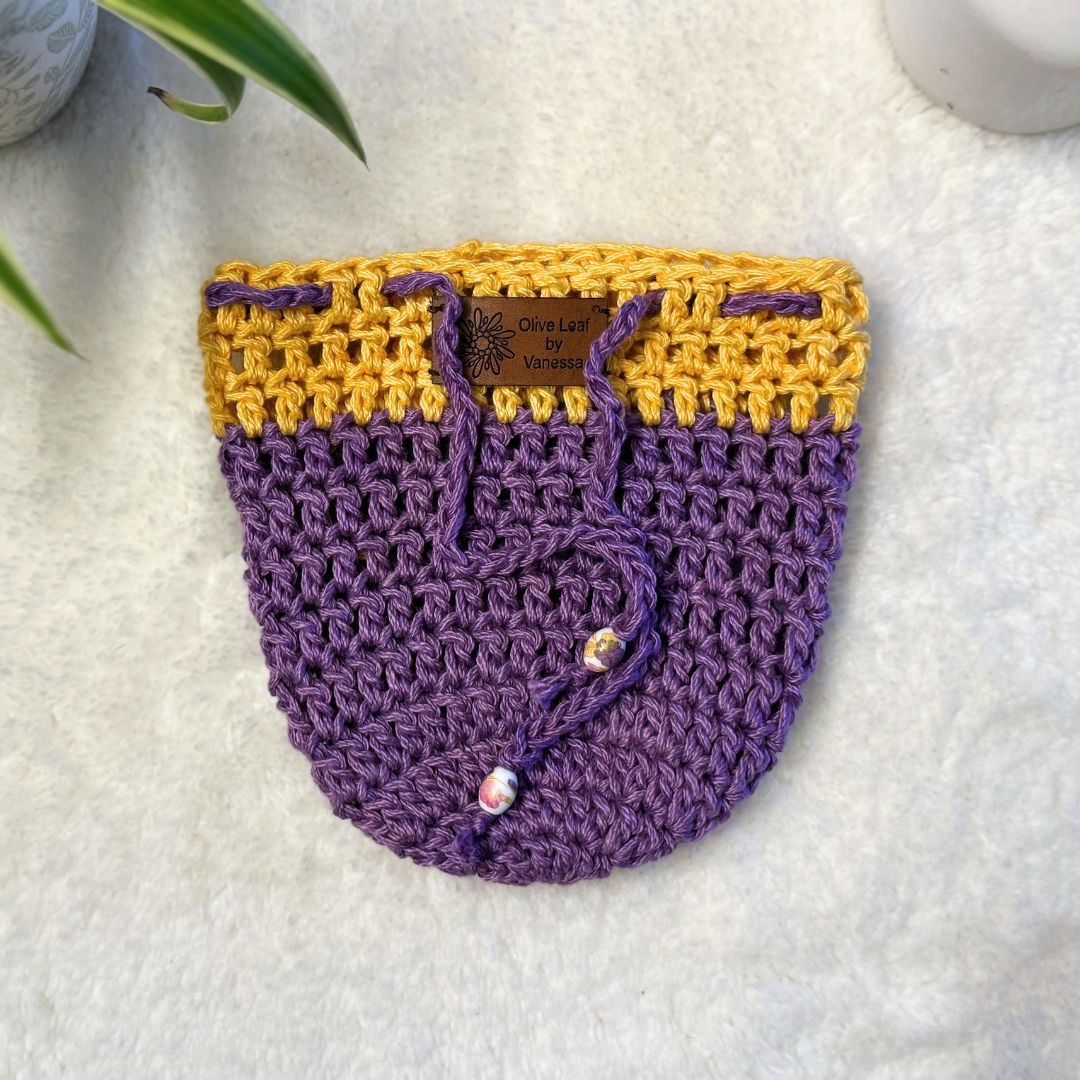 Yellow and purple hand crocheted pot cozy with a drawstring and a faux leather olive leaf label placed on white fur fabric