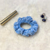 crochet velvet scrunchie in blue placed on a soft fabric with some earrings and a lipstick in the background