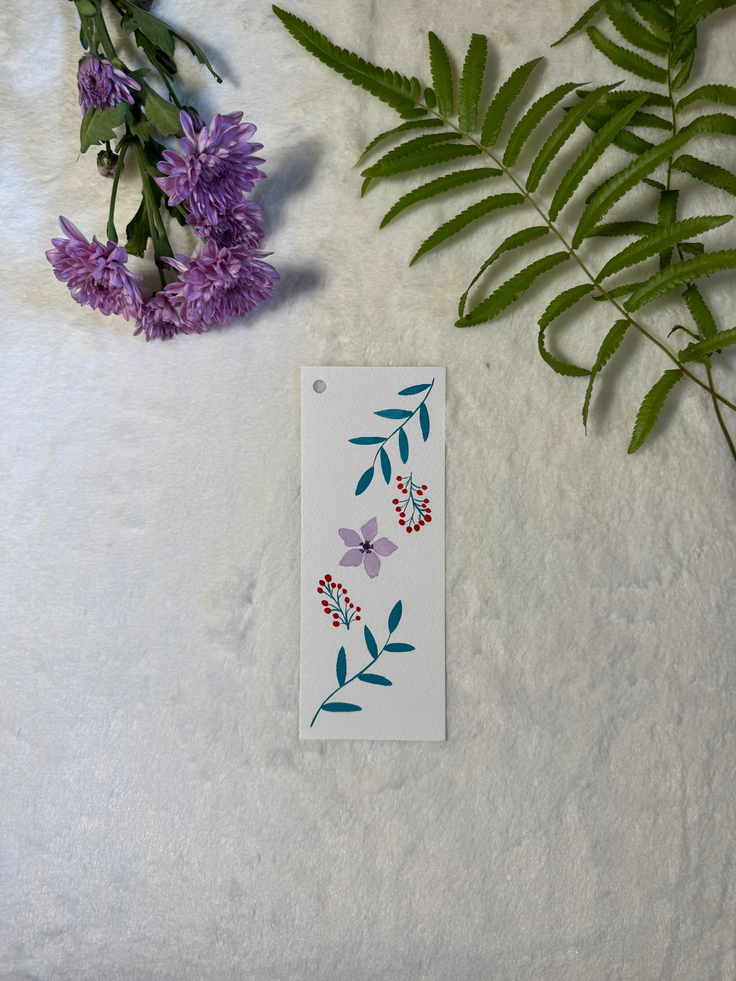 Purple flower and green leaves painted on a bookmark placed on a white fur fabric with fresh flowers and leaves in the background