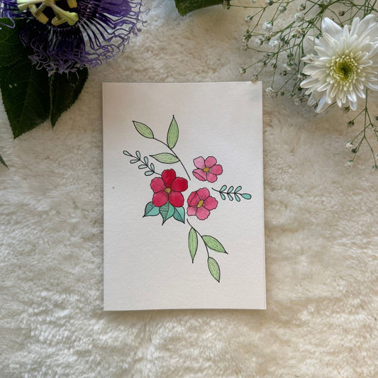 3 flowers in the centre surrounded by leaves hand painted on a greeting card placed on a soft white cloth with flowers in the background