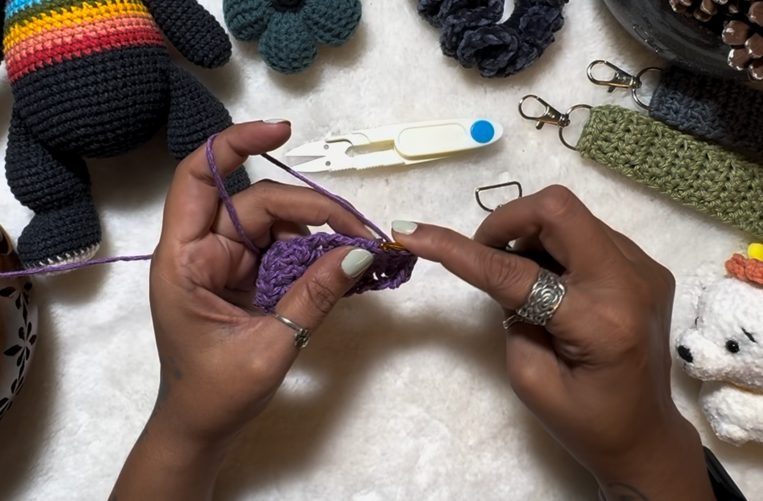 Crochet being done with purple yarn, crochet products in various colours placed on soft fabric