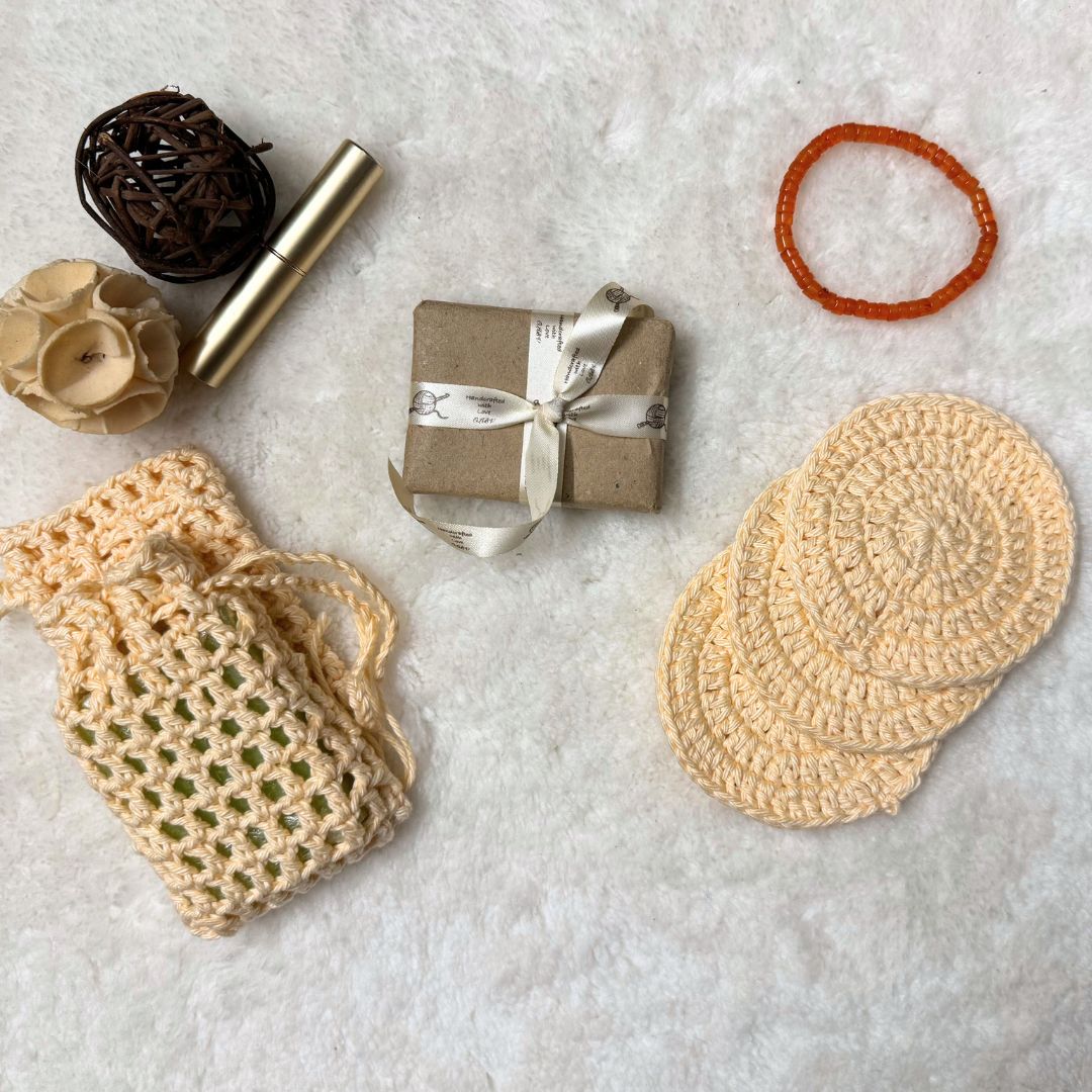 Bath set comprising of 2 soap avers one with a soap inside it, 3 face scrubbies placed on top of each other, a soap wrapped in brown paper and tied with a ribbon and trinkets in the background
