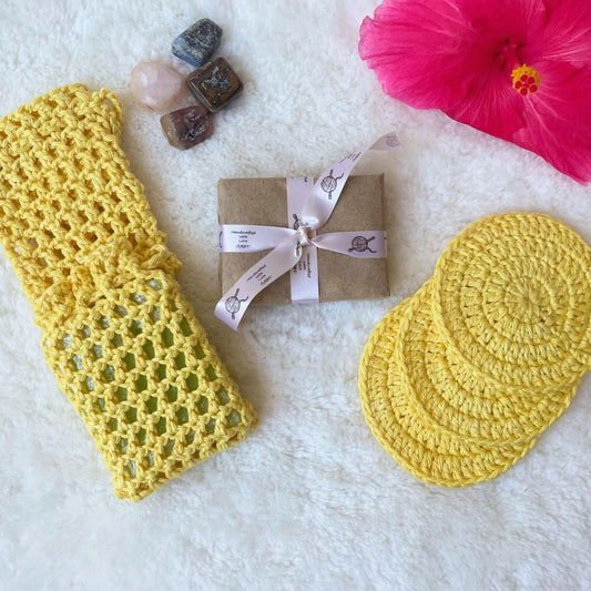Bath set comprising of 2 soap avers one with a soap inside it, 3 face scrubbies placed on top of each other, a soap wrapped in brown paper and tied with a ribbon and trinkets in the background