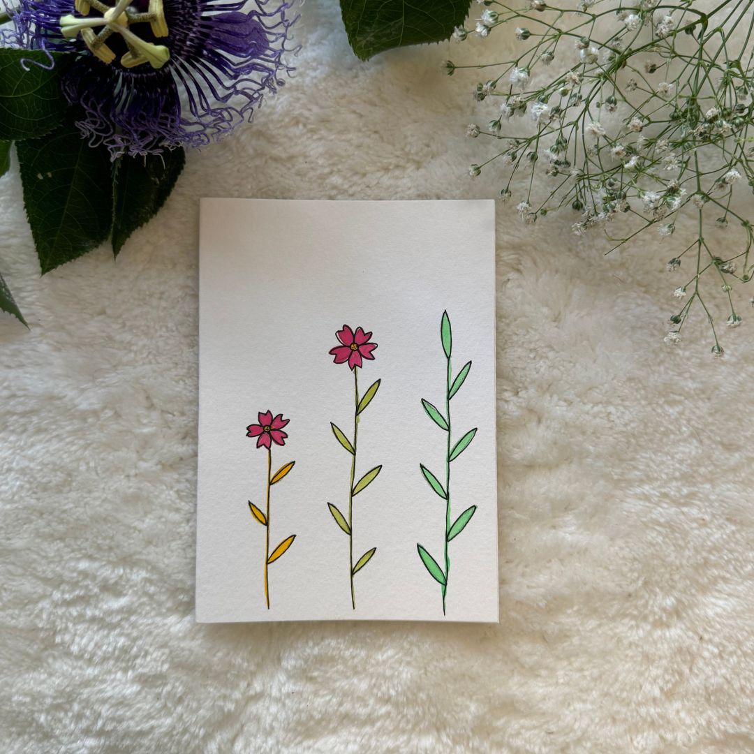 Three single stalk flowers painted on a greeting card placed on a soft white fabric with flowers in the background