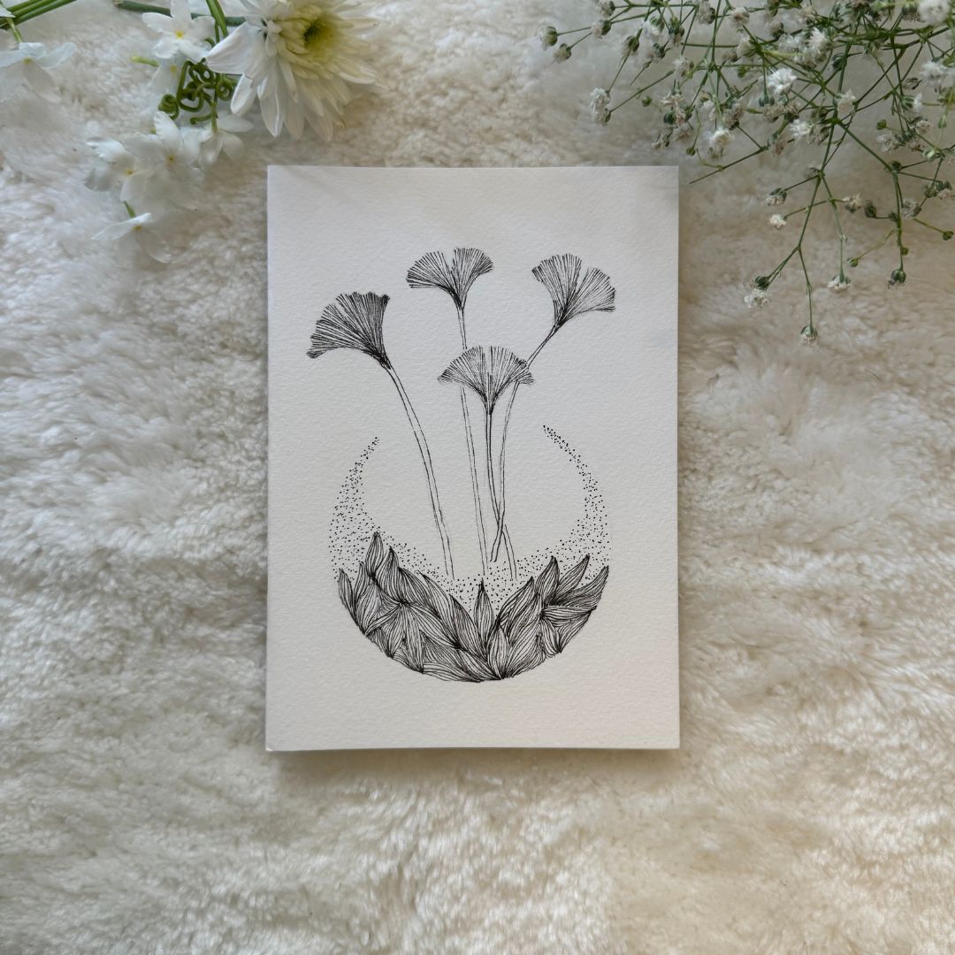 Hand drawn abstract mandala with stripes and dots and ginkgo leaves on a greeting card placed on a soft white fabric with flowers in the background