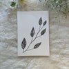 Black and white leaves with lines hand drawn on a greeting card placed on a white soft fabric with flowers in the background