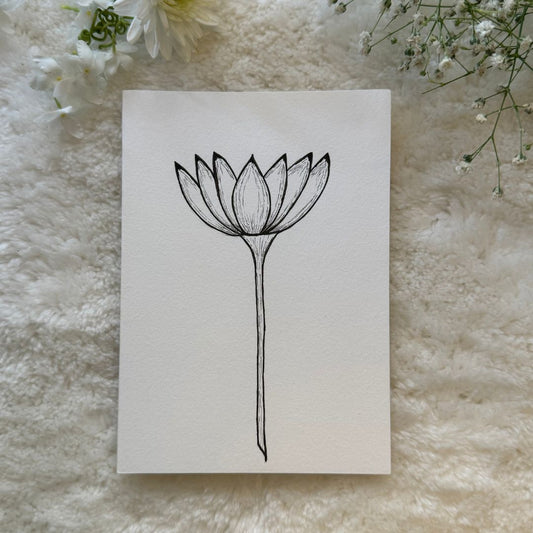 Long stemmed Lotus flower sketched in black and white on a greeting card placed on a soft white cloth with flowers in the background