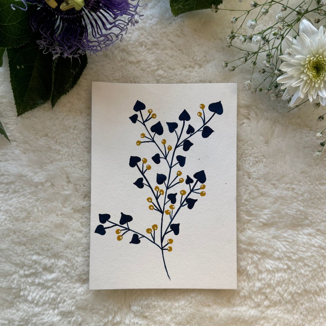 Cherry blossom in gold and navy blue leaves hand painted on a greeting card placed on a soft white cloth with flowers in the background