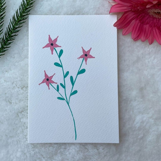 A hand painted greeting card with pink flower stalks painted on a white card placed on a a fur cloth with decorative flowers in the background