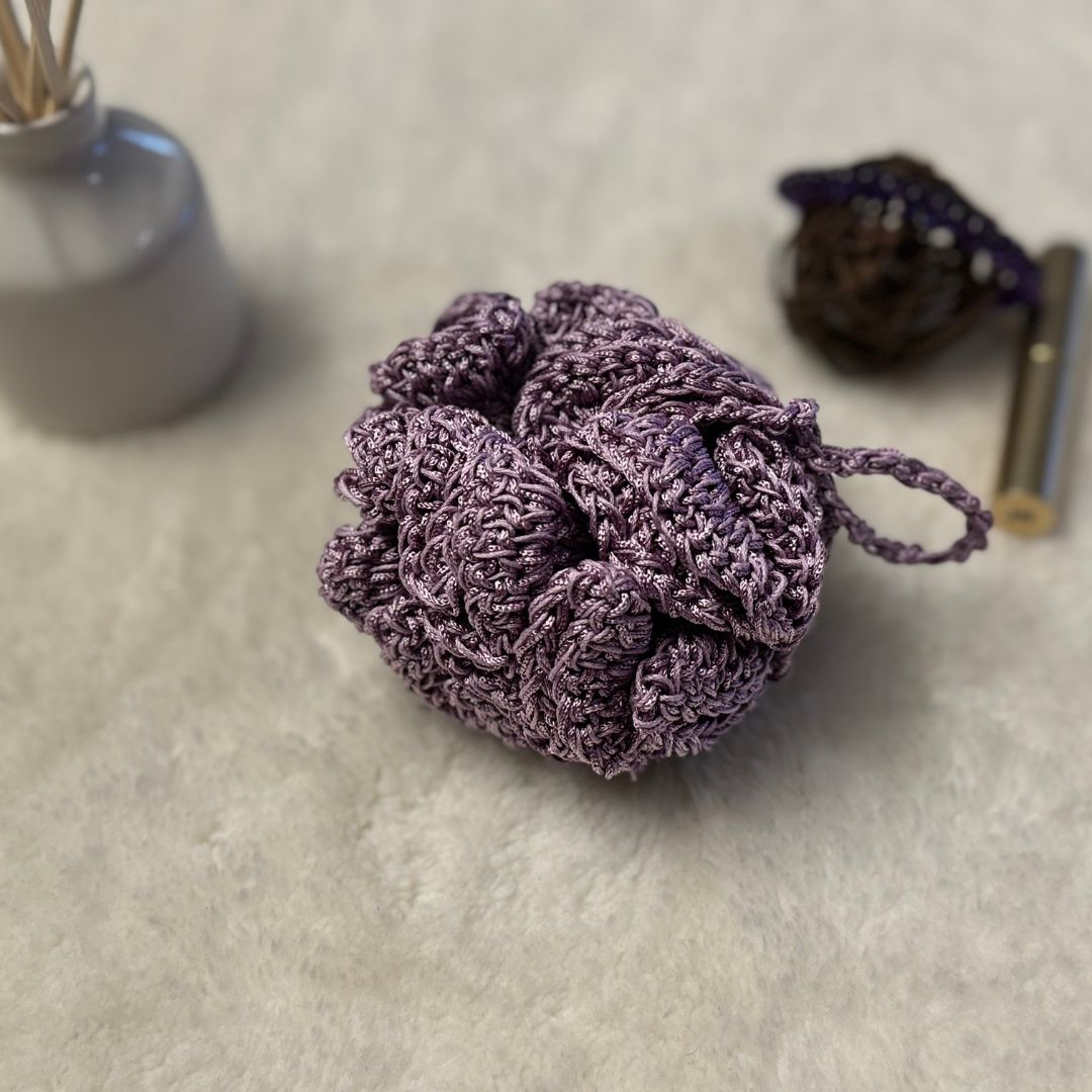 purple crochet loofha placed on a soft white fabric with a few trinkets in the background