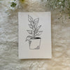 Black and white single stalk pot and plant hand drawn on a greeting card placed on a soft white cloth with lowers in the background