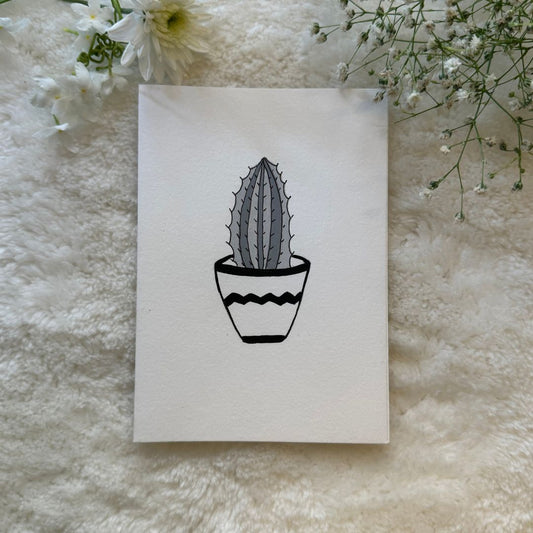 Black and white succulent pot hand painted on a greeting card placed on a soft fabric with flowers in the background