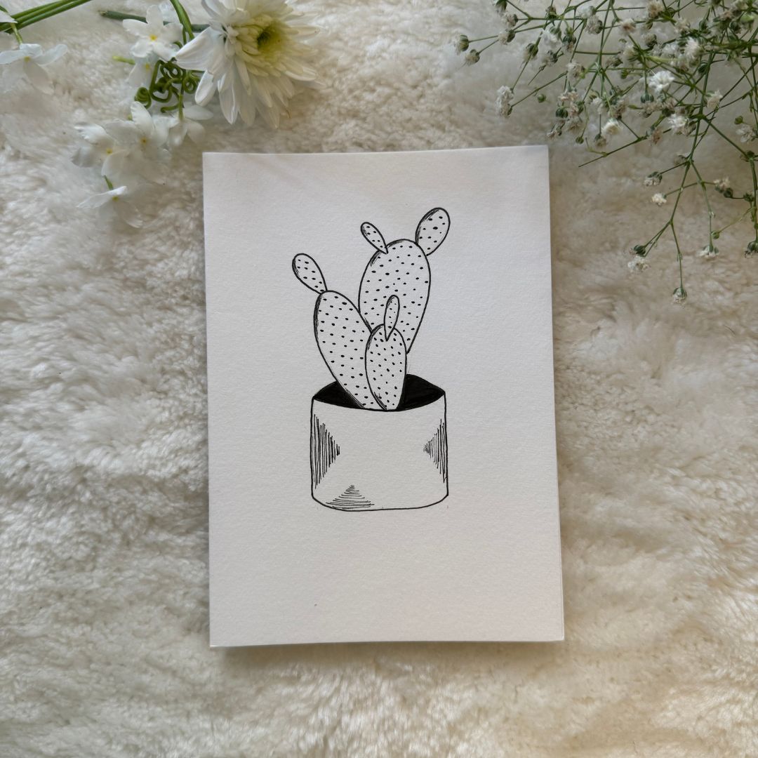 Black and white succulent put with cacti draw on a greeting card placed on a soft white cloth with flowers in the background