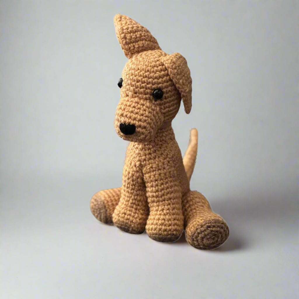 Beige hand crocheted dog sitting with the hind paws spread out and the front paws placed in front with the tail upright 