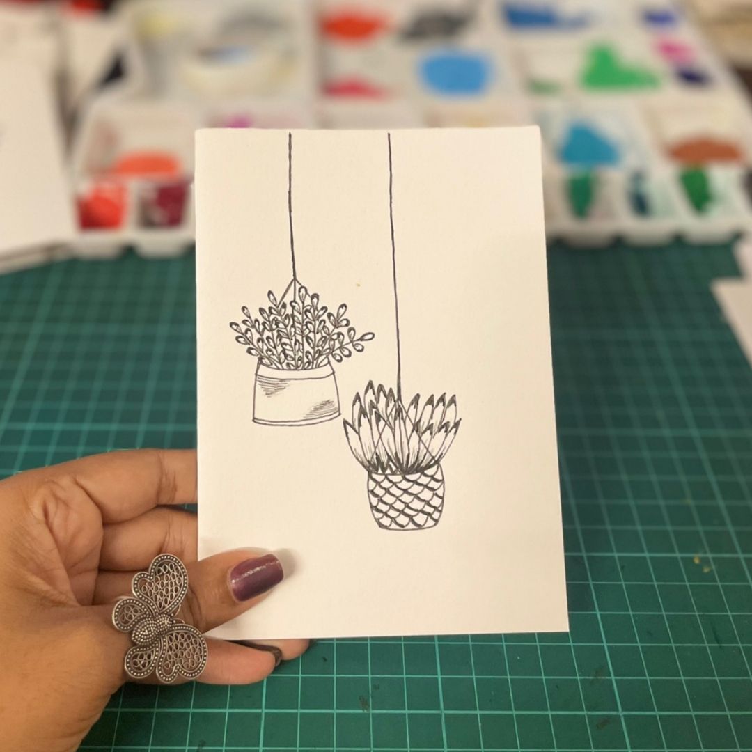 Hand holding a card with black line art of hanging plants on a green cutting mat.