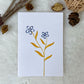 A hand painted greeting card with various flower stalks painted on a white card placed on a a fur cloth with decorative flowers in the background 