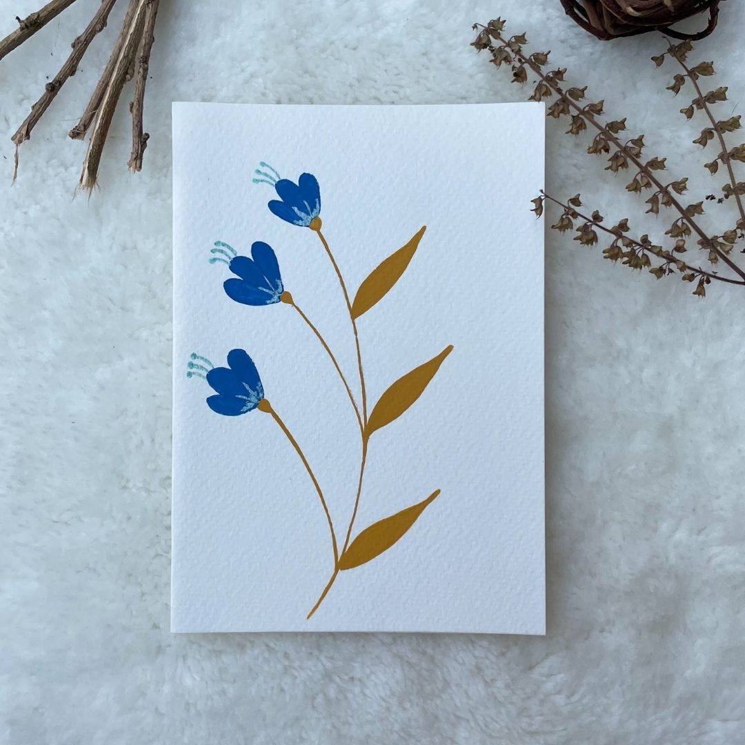 A hand painted greeting card with blue flowers on white card stock placed on a fur surface 
