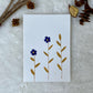 A hand-painted greeting card with blue and mustard gouache illustration of small leaves on a white background, displayed on a fluffy surface with a small branch of leaves to the side.