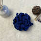 Dark Blue hand crocheted loofha with a hang tie placed on a soft fabric with trinkets in the background