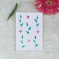 A hand painted greeting card with various flower and stalks painted on a white card placed on a a fur cloth with decorative flowers in the background 