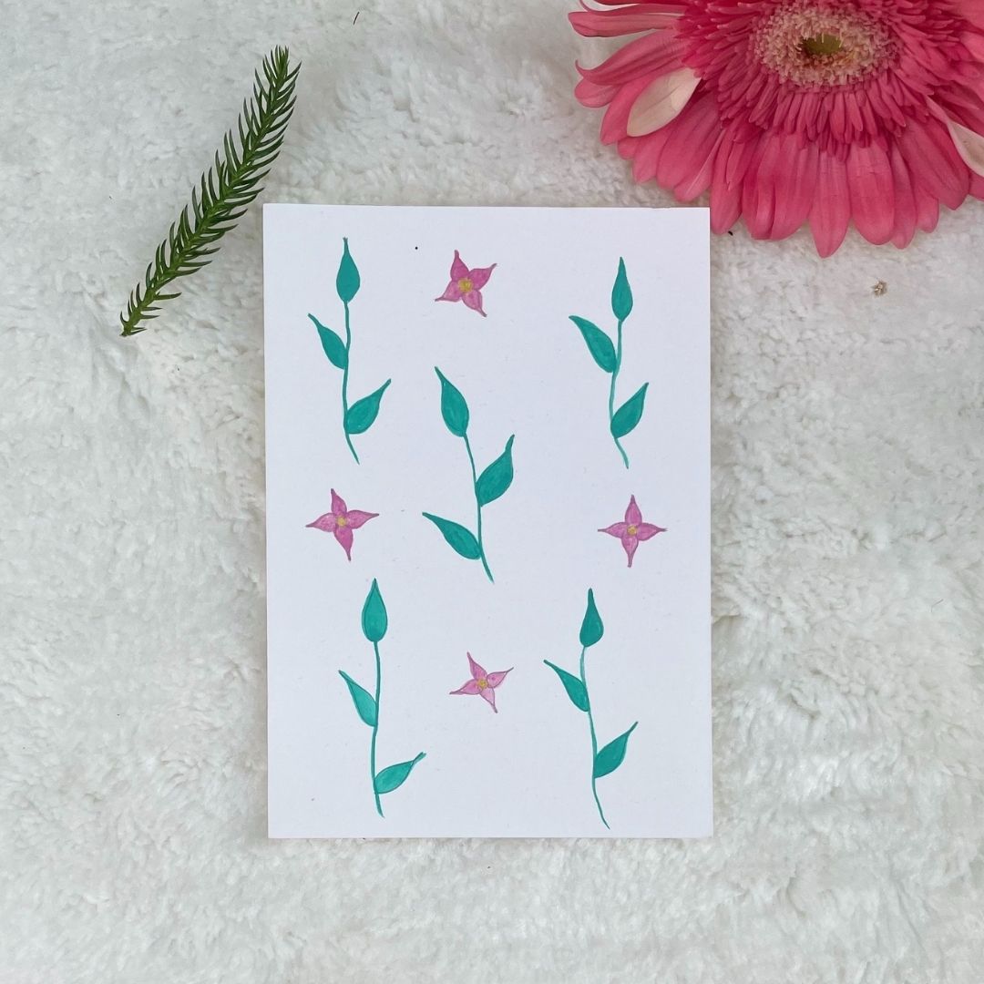 A hand painted greeting card with various flower and stalks painted on a white card placed on a a fur cloth with decorative flowers in the background 