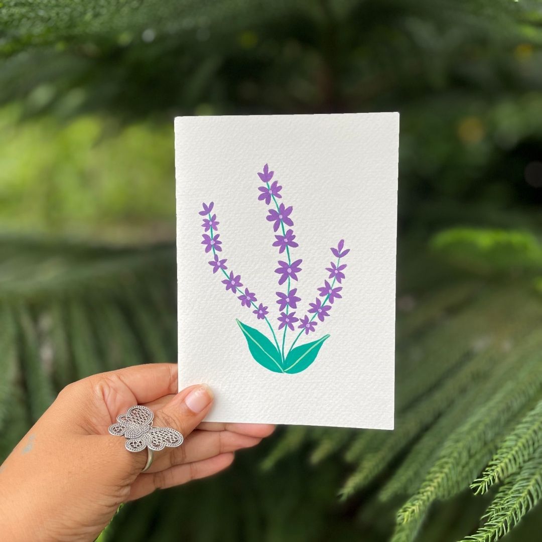 Hand holding a card with a purple flower design against a green leafy background