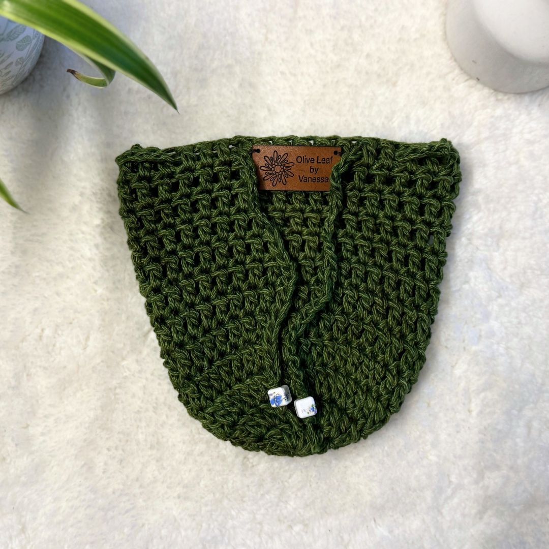 Olive Green hand crocheted pot cozy with a drawstring and a faux leather olive leaf label placed on white fur fabric 