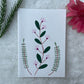 A hand painted greeting card with various flower stalks painted on a white card placed on a a fur cloth with decorative flowers in the background 