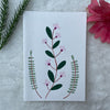 A hand painted greeting card with various flower stalks painted on a white card placed on a a fur cloth with decorative flowers in the background 