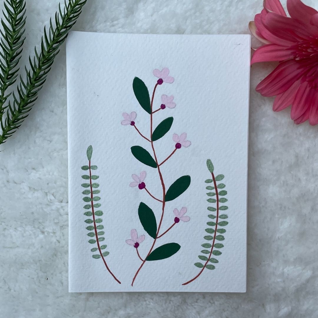 A hand painted greeting card with various flower stalks painted on a white card placed on a a fur cloth with decorative flowers in the background 