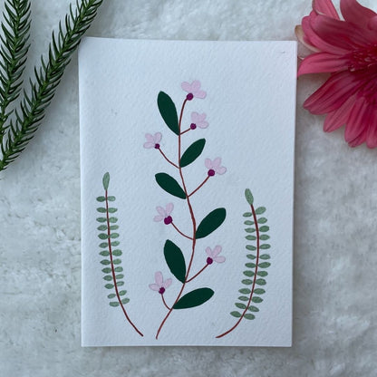 A hand painted greeting card with various flower stalks painted on a white card placed on a a fur cloth with decorative flowers in the background 