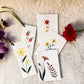A set of white botanical art cards featuring hand-painted flowers and leaves, arranged on a textured cream fabric alongside purple orchids and a red flower.