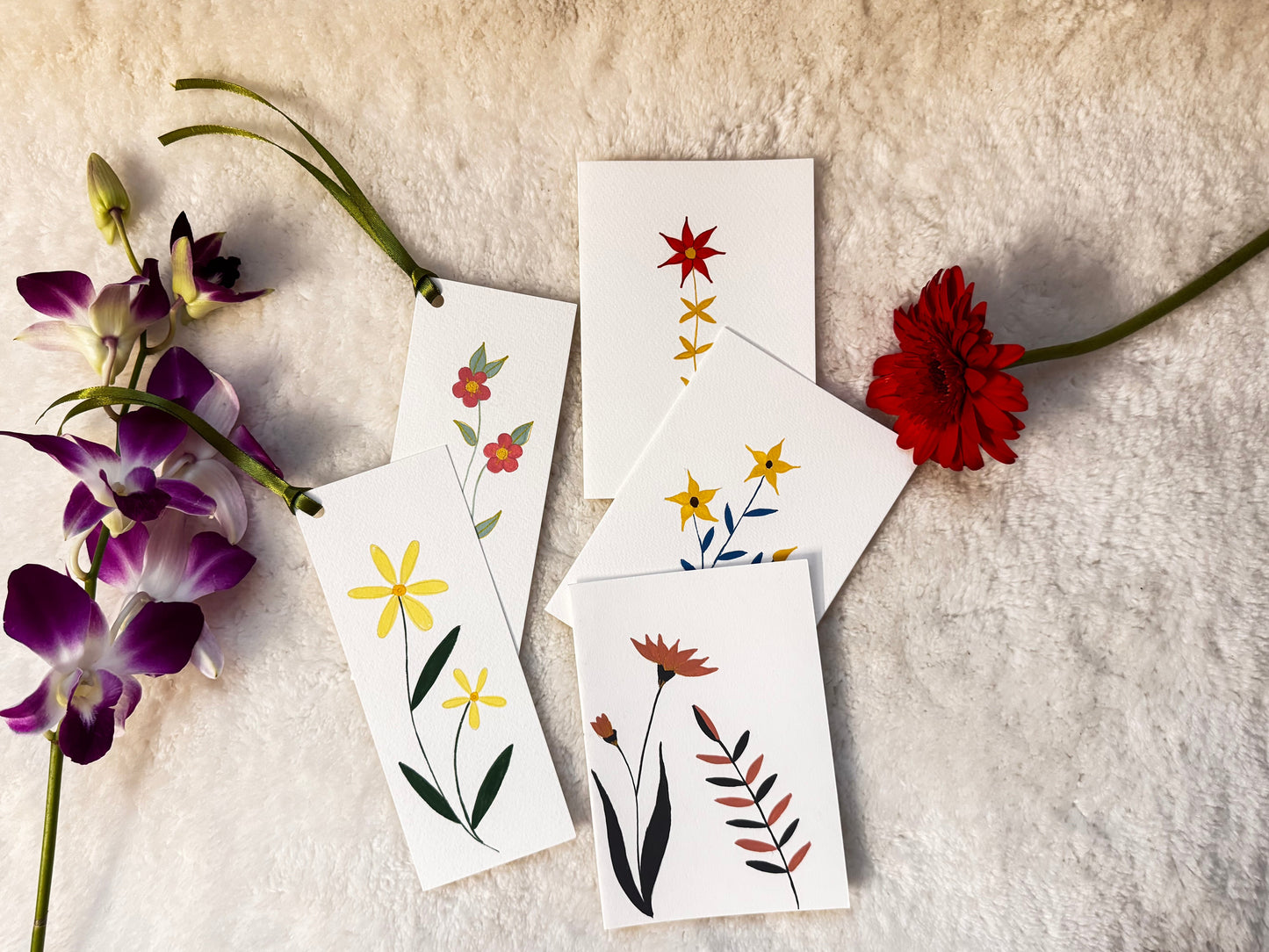 A set of white botanical art cards featuring hand-painted flowers and leaves, arranged on a textured cream fabric alongside purple orchids and a red flower.