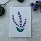 A hand painted greeting card with various flower stalks painted on a white card placed on a a fur cloth with decorative flowers in the background 