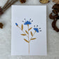 A hand painted greeting card with various flower stalks painted on a white card placed on a a fur cloth with decorative flowers in the background 