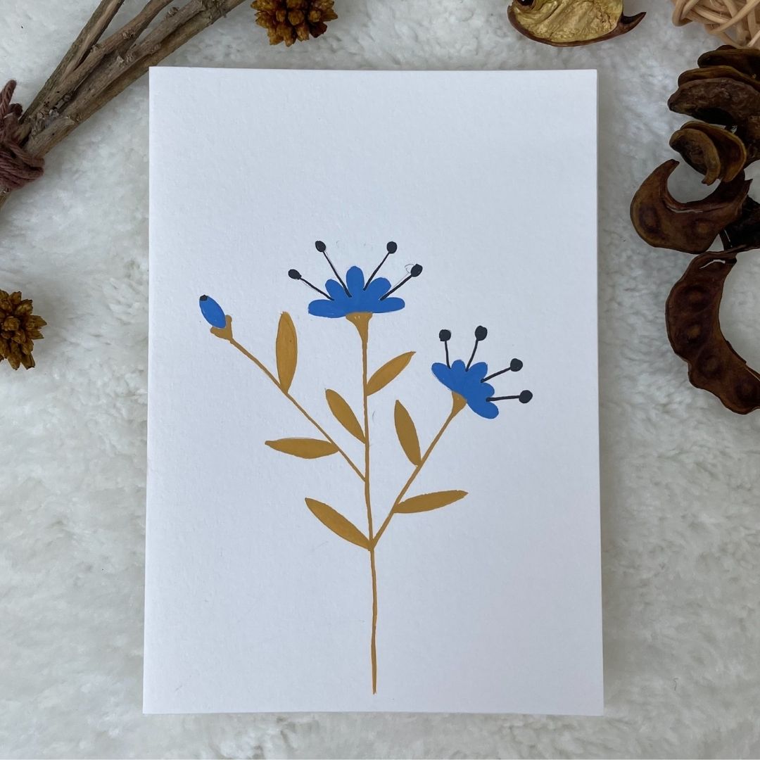 A hand painted greeting card with various flower stalks painted on a white card placed on a a fur cloth with decorative flowers in the background 