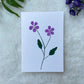 A hand painted greeting card with purple flower stalks painted on a white card placed on a a fur cloth with decorative flowers in the background 