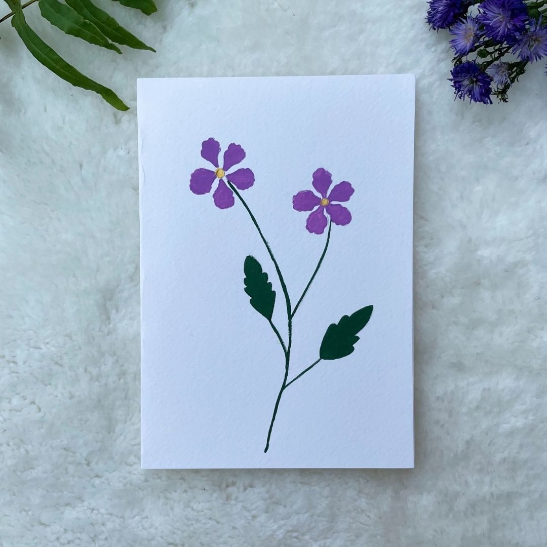 A hand painted greeting card with purple flower stalks painted on a white card placed on a a fur cloth with decorative flowers in the background 