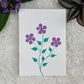 A hand painted greeting card with Purple flowers on a white card placed on a fur background 