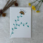 A hand painted greeting card with a yellow flower stalk painted on a white card placed on a a fur cloth with decorative flowers in the background 