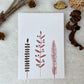 A hand painted greeting card with various flower stalks painted on a white card placed on a a fur cloth with decorative flowers in the background 