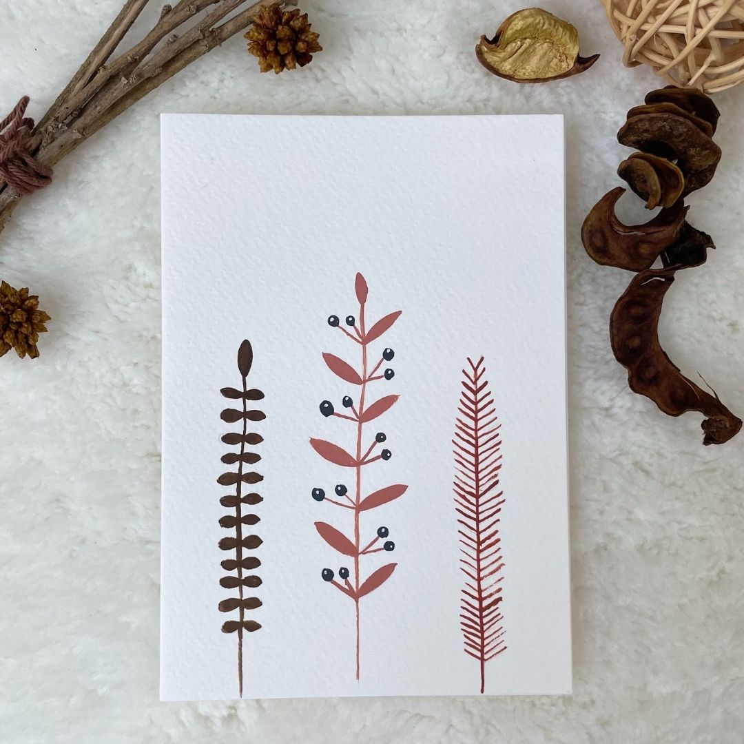 A hand painted greeting card with various flower stalks painted on a white card placed on a a fur cloth with decorative flowers in the background 
